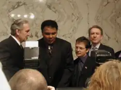 Michael J. Fox and Muhammad Ali are seen speaking to reporters, with the marble walls of the U.S. Senate behind them