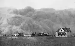 A dust storm approaches a collection of houses, dwarfing them in height.