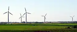 A car drives past 4 wind turbines in a field, with more on the horizon