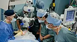 A surgical team is gathered around the patient in an operating theatre. the surgeon and two learners are observing the procedure through a surgical microscope suspended above the patient's eye.
