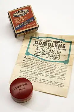 A jar of ointment, with a box and a poster. The box has the words "Domolene Brand Stops all skin troubles rashes and irritation The miracle ointment".