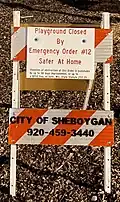 A sign upon a traffic barricade in front of a playground in Sheboygan notifies the public that the equipment is closed due to Evers's stay-at-home order, and of the consequences for violating it.