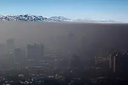 Dense, smokey air lays over a cityscape like a blanket. In the distance, a mountain range and clear blue sky can be seen.