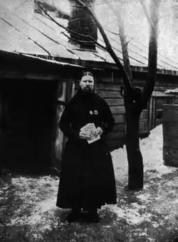 An old man with a beard and long hair wearing black standing in front of a wooden house.