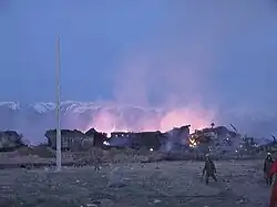 In twilight against snow capped mountains, two firefighters walk through a field flanked by burning rubble of the village