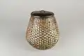 A small, squat water jar with a solid black lid and a brown and cream glaze, shaped into the densely-woven texture of a woven bamboo basket.