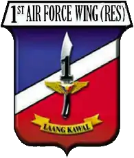 A shield-style crest primarily in red and blue, divided with a diagonal white bar. The centre contains an symbolic military knife with the number '1' covering its blade. The crest is headed by '1st Air Force Wing (Res)' in all-upper-case letters. Below the knife is a yellow banner for its motto containing the phrase 'Laang Kawal'.