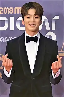 Kim Min-kyu wearing a tuxedo on a red carpet, smiling and holding up two finger hearts