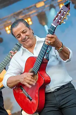 Jalloul Bouchikhi wearing a white shirt and dark pants, playing a red acoustic guitar onstage