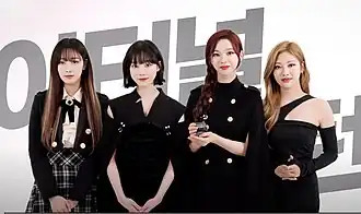 Four women dressed in black outfits, standing together in front of a background with partially visible Korean text with one of them holding a small trophy.