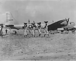 A black and white photograph. An aircraft is in the background with soldiers and labourers working around it. Five women are in the foreground carrying construction materials in baskets