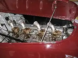 Close-up of a straight-eight engine installed in the front of a racing car, as seen through the right side of the bonnet, which is hinged in the centre and is held open with a metal rod. There are four downdraft carburettors on the near side of the engine, these have intake stacks angled backward and toward the engine. The exhaust manifold is visible on the same side of the engine as the carburettors.