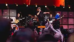 Five men perform on stage: The rhythm guitarist looking at his guitar, drummer behind his kit, lead singer in the middle, bass guitarist turned to his right and lead guitarist in school boy costume.