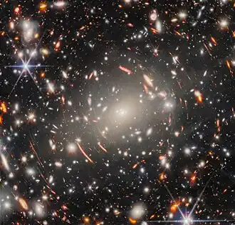 A field of galaxies in space, dominated by an enormous, bright-white elliptical galaxy that is the core of a massive galaxy cluster. Many other elliptical galaxies can be seen around it. Also around it are short, curved, glowing red lines, which are images of distant background galaxies magnified and warped by gravitational lensing. A couple of foreground stars appear large and bright with long spikes around them.