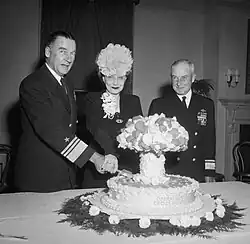 A man in naval uniform and a woman wearing a hat cut into a cake labeled Operation Crossroads, and shaped like a mushroom cloud, while another naval officer looks on.