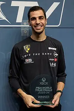 A portrait photo of Ali Farag where he is standing against the side wall of the squash court, holding the winner's trophy in his hands in front of his waist.