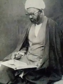 Hussein Bakhsh Jara sits wearing glasses with open book and pen in hand