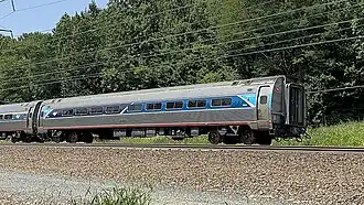 A silver railroad car with a blue stripe over the windows. At both ends, an angular light-blue shape covers the stripe.