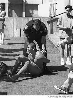 A nude man with a shaved head seated on the ground being arrested by police officers