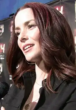 A 31-year-old woman with long, brown hair, smiling with her head tilted at someone holding a microphone to the left of the camera.