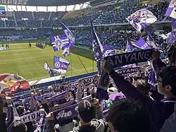 In the background, teams line up on a football pitch in a large stadium. In the foreground, fans of FC Anyang hold up purple scarves and wave flags in support of their team.