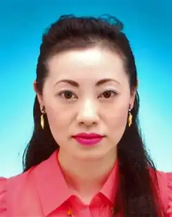 A portrait of an Asian woman with long hair, arranged in front of body on one side, wearing a pink shirt and yellow-orange earrings, looking straight at the camera.
