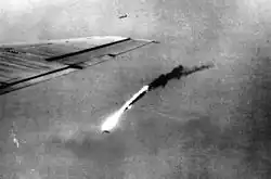 Black and white photo of flaming wreckage falling towards the ground. The wing of a plane is visible at the left-hand side of the photo.