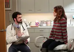 A woman sits in a doctor's office whilst a bearded doctor talks to her in a chair