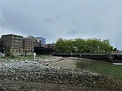 A stony beach overlooking several piers, brick buildings, and some trees
