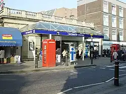 A single storey pale brick building topped with stone railing. Above an entrance is a canopy, around which are rectanglar blue signs reading "BAYSWATER" and "BAYSWATER STATION". People are walking in the street.