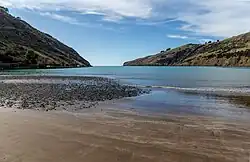 A perspective looking out from a sandy, brownish-grey beach (covered in to the dark pebbles to the left and smooth to the right) toward the somewhat-distant mouth of the bay, which is flanked on either side by peninsulas. The left is steep and lightly forested, while the right is shallow and bald except for grass. Very weak waves wash up onto the shore.