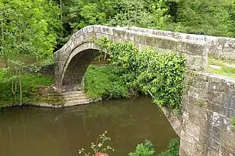 An arched stone bridge, spanning a narrow body of still water, with trees on the far side of the river