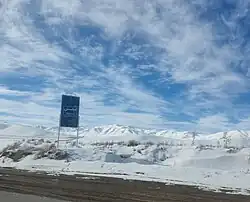 A clear road surrounded by snow, with snowy mountains in the distance. There is a road sign with the town name in English and Arabic