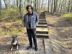 Performing and recording artist Bill Nace standing at the base of a trail surrounded by trees and next to a dog