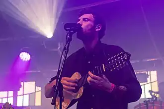 Blanco White wearing a dark shirt, standing onstage, playing a charango and singing into a microphone