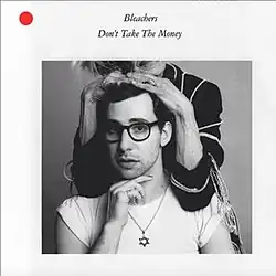 A picture of Jack Antonoff wearing a white T-shirt and a Star of David necklace. He rests his hand on his chin. An elderly woman places her hands on his hair, with her face out of frame.