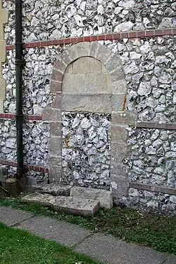 Part of a flint wall with red-brick bands, in which a tall arch-shaped blocked entry is set. It has flints and stone blocks, and is surrounded by stones of different sizes.
