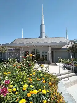 The front of the building is seen with a bunch flowers in the bottom left corner. A concrete path leads to the temple.