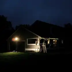A photograph of a person laying down in front of a house, emphasized by a floodlight. Two skeletons are hanging lights on the house.