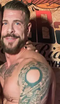 Rocco Steele standing shirtless next to Boomer Banks. Steele is a middle-aged white man with short hair, facial hair, and blue eyes. He has a muscular figure and torso and upper-arm tattos.