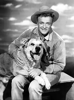 A black and white photograph of Brian Keith in a cowboy costume sitting with a large dog sitting in his lap.