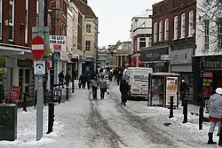 Street scene with pedestrians and vehicles in road lined with shops. There is snow on the ground.