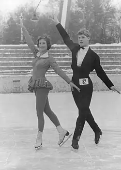An image of Margit Senf and Peter Göbel skating side-by-side. They both have their right hand pointed upwards and their left hand pointed downwards with their feet crossed.