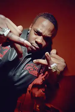 A picture of a man displaying the jewelry on his fingers to the camera. He wears a red-black jacket and stands in front of a red background.