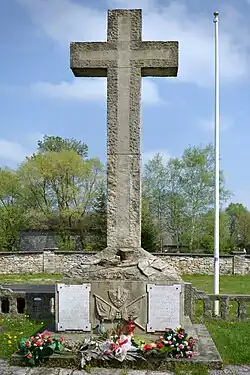 A photograph of the memorial, which consists of a large crucifix made of stone with two inscribed plaques at the bottom. Some flowers have been placed in front of it.