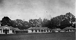 Black and white photo of a long one-story building, behind which is a row of trees