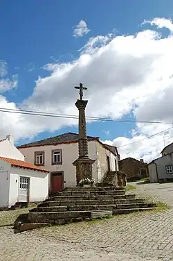 A monument in the form of a pillar topped with a crucifix on a steep cobblestone street. Two white buildings with tile roofs stand behind it.