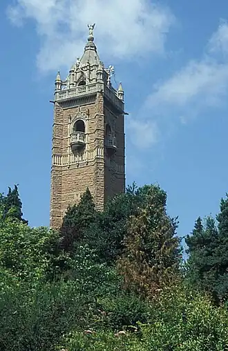 An ornate brick tower surrounded by trees. The tower has balconies and is surmounted by a pitched roof with an ornate figure at the apex.