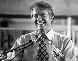 Jimmy Carter stands smiling in a shirt and tie in front of microphones.