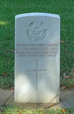 A white grave stone marker. The stone contains an insignia at the top with a crown, flying eagle and banners, followed by text of the person and a Christian cross.